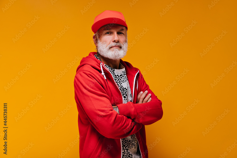 Portrait of man in red cap and hoodie on orange background. Modern guy with grey beard in bright clothes looking into camera.. Portrait of man in red cap and hoodie on orange background. Modern guy with grey beard in bright clothes looking into camera..