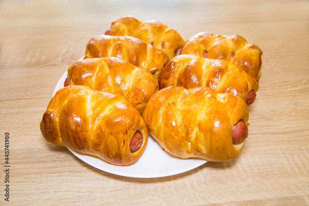 Sausages in a delicious yellow dough on a white round plate on a wooden board background. Bakery products national traditional dishes and pastries. Sausages in a delicious yellow dough on a white round plate on a wooden board background. Bakery products national traditional dishes and pastries.