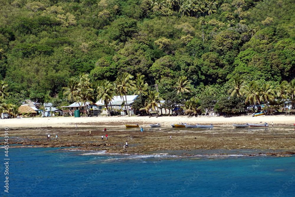 People on a white sand beach near a village on a bay below a jungle in Fiji