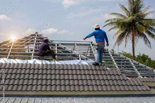 Roof repair, worker with white gloves replacing gray tiles or shingles on house with blue sky as background and copy space, Roofing - construction worker standing on a roof covering it with tiles.