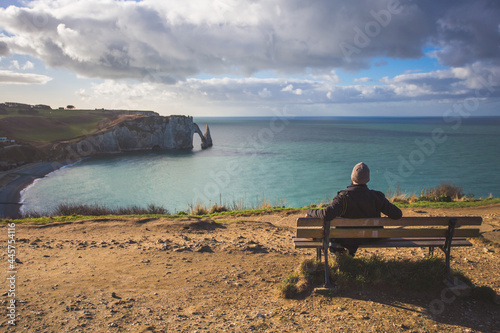Fotografi Tourist sitting on a bench admiring the seascape Etretat Aval cliff, rocks and natural arch landmark and blue ocean