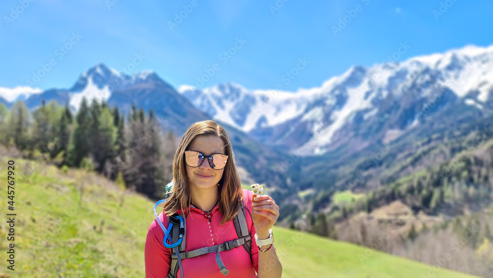 A woman holding daisies in her hand with panoramic view on Baeren Valley in Austrian Alps. The highest peaks are sonw-capped. Lush green pasture in front. Clear and sunny day. High mountain chains.