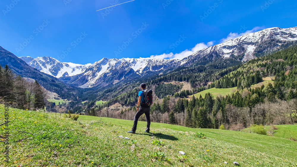 A man with a big hiking backpack enjoying the panoramic view on Baeren Valley in Austrian Alps. The highest peaks are sonw-capped. Lush green pasture. Clear and sunny day. High mountain chains.