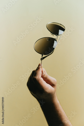 Cuadro en lienzo Hand grabbing a pair of dark sunglasses over a pastel yellow background, style a