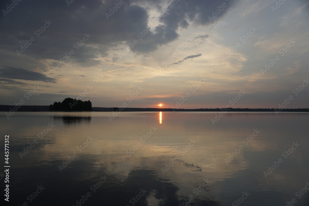 Obraz premium Evening on the lake in summer in July. The sun and sky are reflected on the calm surface of the water. Soft light and bright colors. Sunset and pacification