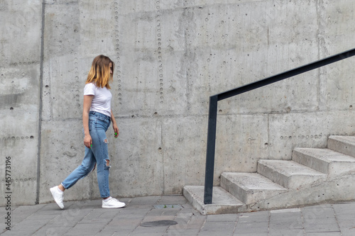 A young woman in casual clothing walks along the sidewalk past a concrete wall.