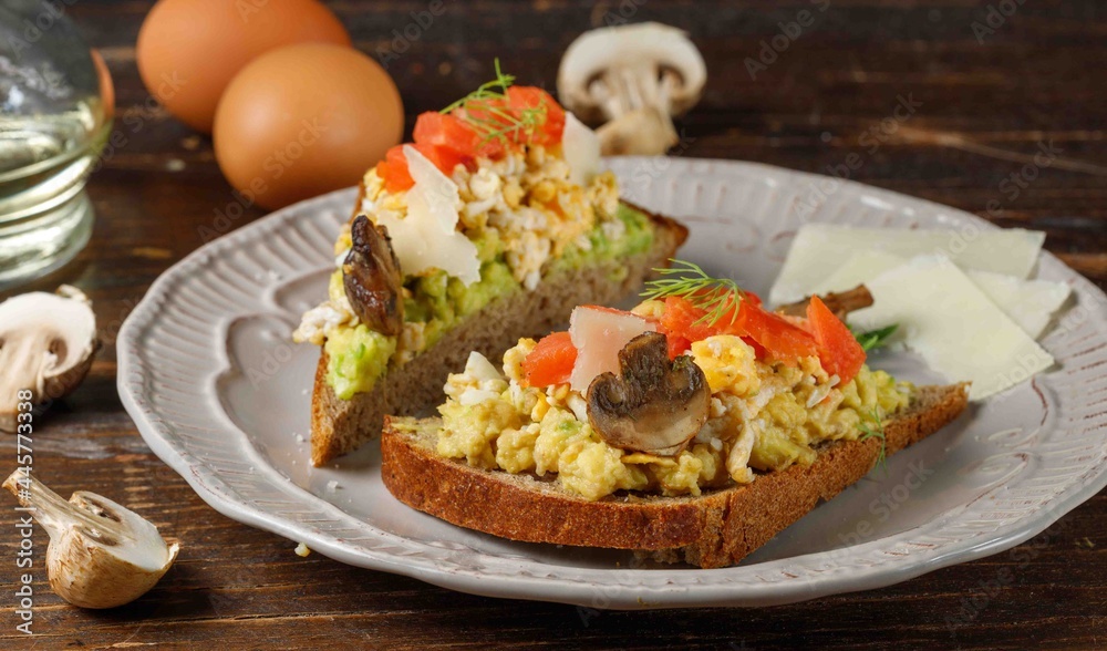 Two toasts with guacamole, scrambled eggs, salmon and mushrooms. On rye bread. Close-up, on a gray plate. Dark wooden background. Two toasts with guacamole, scrambled eggs, salmon and mushrooms. On rye bread. Close-up, on a gray plate. Dark wooden background.