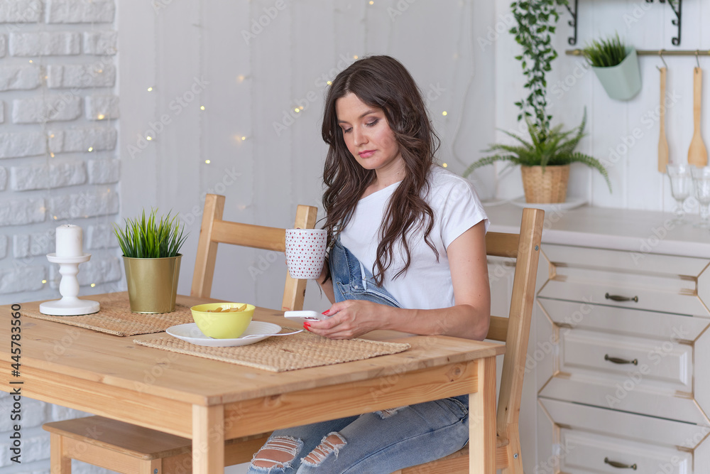 female smiling using mobile phone holding coffee at home in kitchen. ordering food through online stores. buying food through an online application