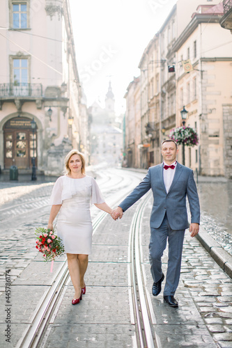 Cheerful shot of lovely middle aged married couple posing on camera, while walking holding hands on tram track on pavement road, in old ancient city. Family, love and people concept © sofiko14