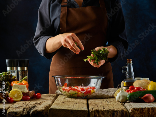 The chef in a brown apron adds lettuce to the Greek salad. Many bright ingredients for Greek salad are on the table side by side. A beautiful still life. Bright colors. Dark blue background.