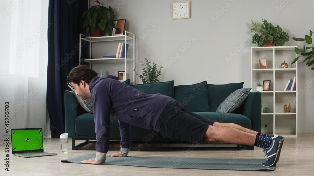 Young man doing push-ups from the floor in living room, using video fitness lesson on laptop computer, green screen, chroma key. Remote online sport during coronavirus quarantine isolation at home. Young man doing push-ups from the floor in living room, using video fitness lesson on laptop computer, green screen, chroma key. Remote online sport during coronavirus quarantine isolation at home.