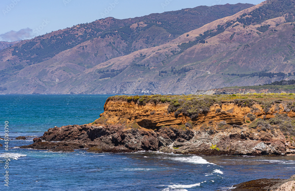 San Simeon, CA, USA - June 8, 2021: Pacific Ocean coastline North of town. Clsoeup of Red rock cliffs at deep blue water near Ragged Point with Los Padres mountains in back.