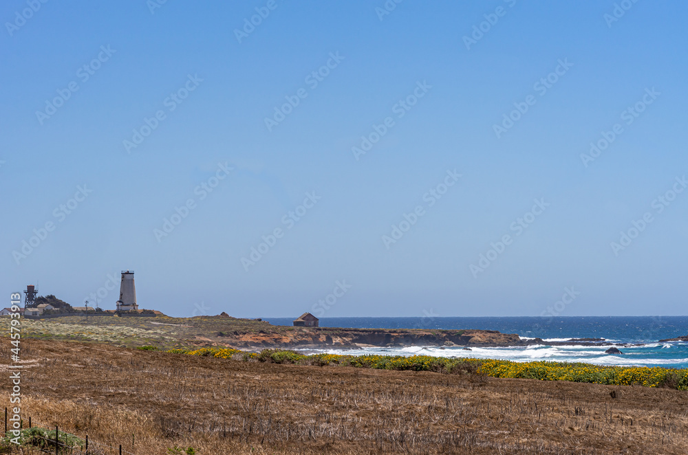 San Simeon, CA, USA - June 8, 2021: Pacific Ocean coastline. Wide landscape of Point Piedras Blancas with its lighthouse near white surf of deep blue water under light blue sky.