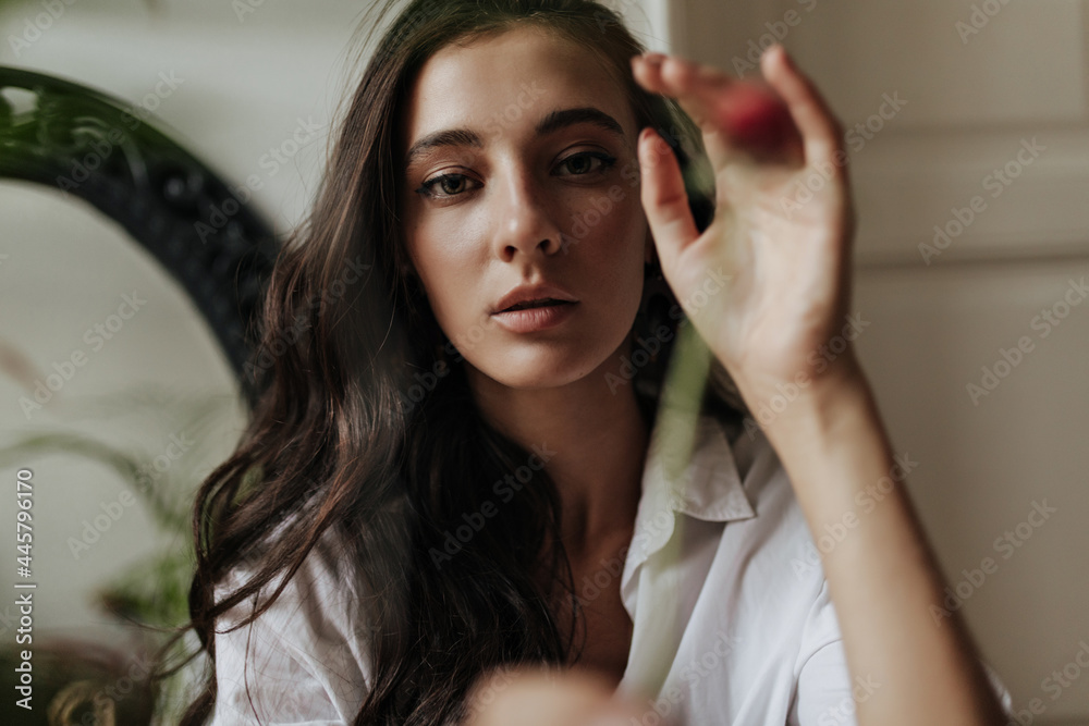 Fototapeta premium Charming modern girl with tanned skin and curly brunette hairstyle in stylish white shirt looking into camera in apartment..