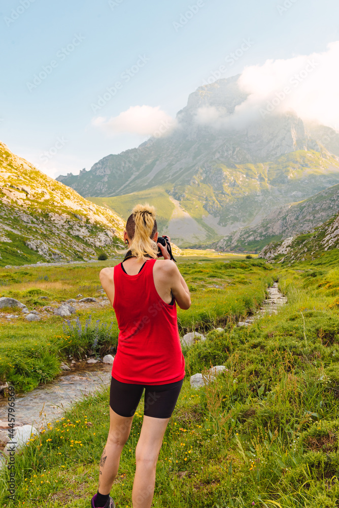 Caucasian young woman from behind next to a river and near a mountain taking pictures of the landscape during a trip. Picos de Europa National Park.