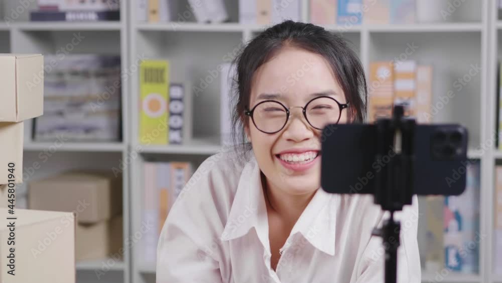 Happy young Asian woman wearing glasses and sitting to do live on social media at home, setting camera on a table, Women doing business selling online, online sales channel, woking at home Happy young Asian woman wearing glasses and sitting to do live on social media at home, setting camera on a table, Women doing business selling online, online sales channel, woking at home