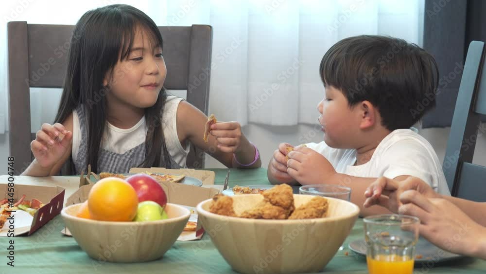 Happy Asian family parents with little daughter and son eating fried chicken and pizza for dinner together. Father and mother with child boy and girl enjoy eating and sharing a meal together at home. Happy Asian family parents with little daughter and son eating fried chicken and pizza for dinner together. Father and mother with child boy and girl enjoy eating and sharing a meal together at home.