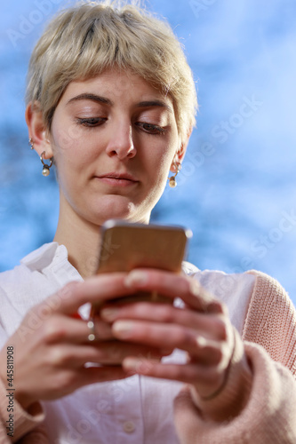 Shot from below of young adult woman typing text message on smartphone. Selective focus on face. In the background light blue sky. Vertical image.