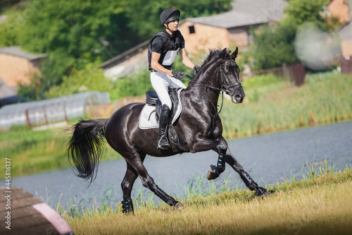 portrait of rider man and black stallion horse galloping fast near lake during eventing cross country competition in summer