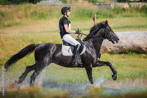 rider man galloping fast in water pond on black stallion horse during eventing cross country competition in summer