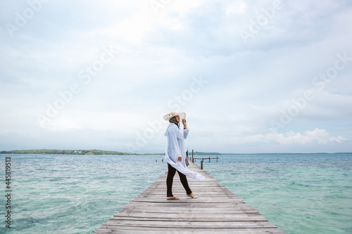 Woman tourist in white dress and summer hat posing on the wooden dock with seascape background