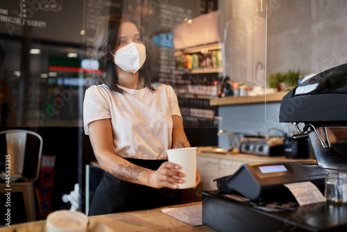 Female barista at cafe giving coffee cup through protective plastic screen wearing mask
