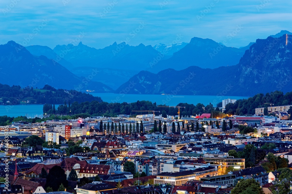 Aerial panorama of beautiful Lucerne City by lakeside at blue dusk with majestic mountains in background and lights of houses under evening twilight before nightfall, in Switzerland, Europe
