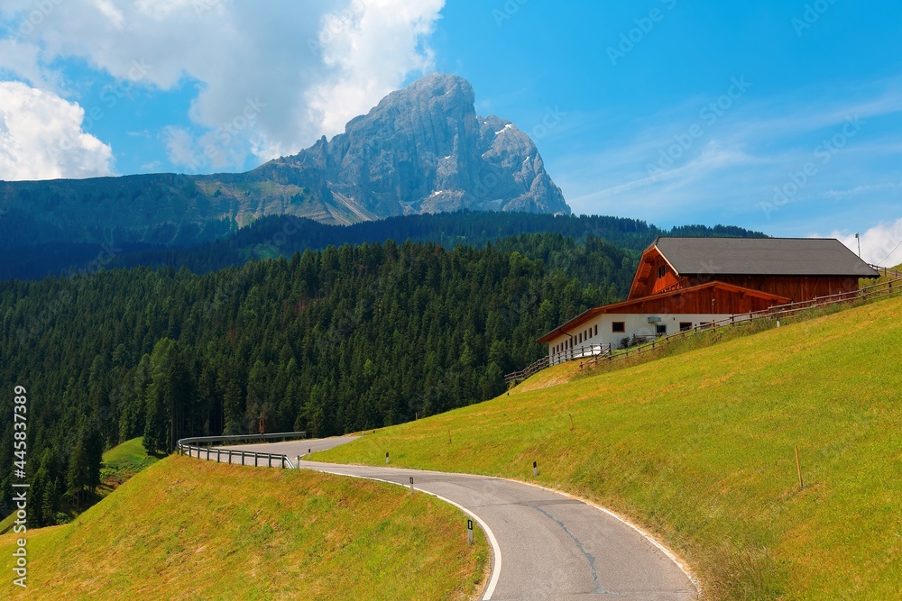 Summer scenery of beautiful Dolomites with majestic Peitlerkofel ( Sass de Putia ) Mountain in background & a farmhouse by a curvy highway winding through green grassy hillside in South Tyrol, Italy