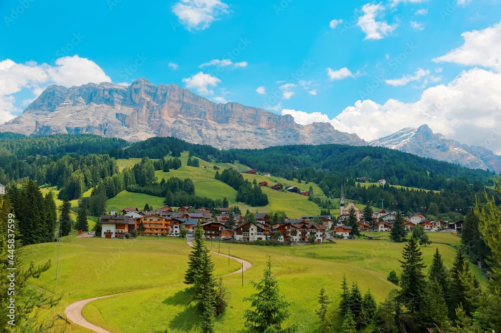 Summer scenery of a beautiful village at foothills of rugged Sasso di Santa Croce & a country road winding in the green grassy valley in Val Badia, Alta Badia, Dolomiti, Trentino, South Tyrol, Italy