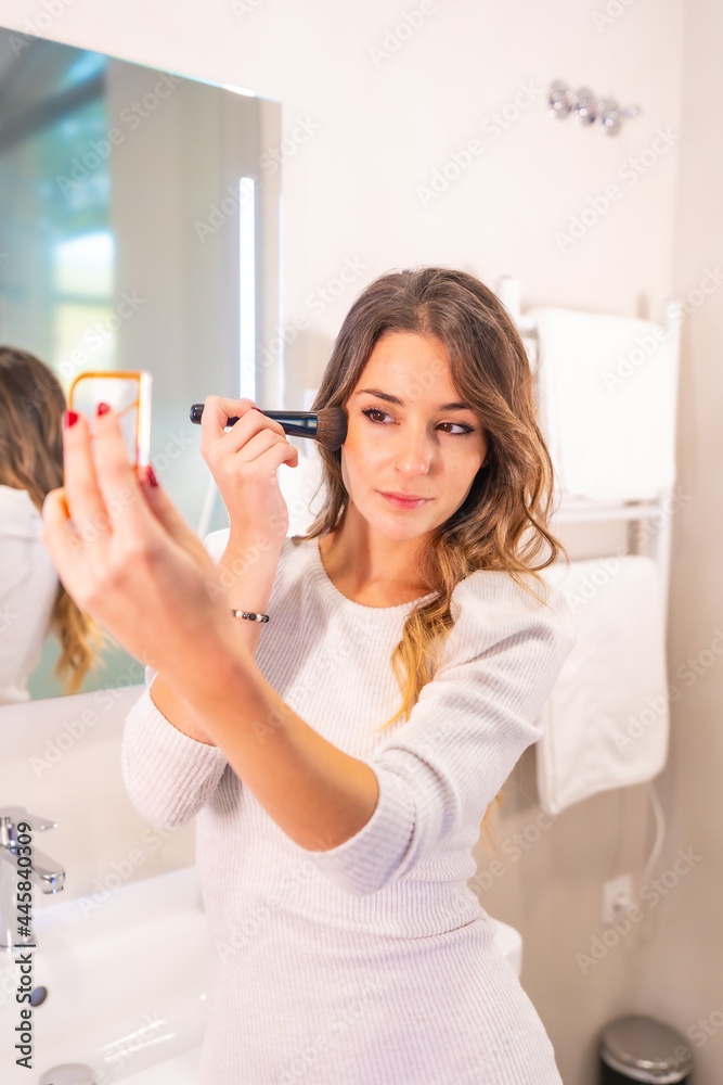 Caucasian woman putting on makeup in bathroom looking in mirror, vertical photo
