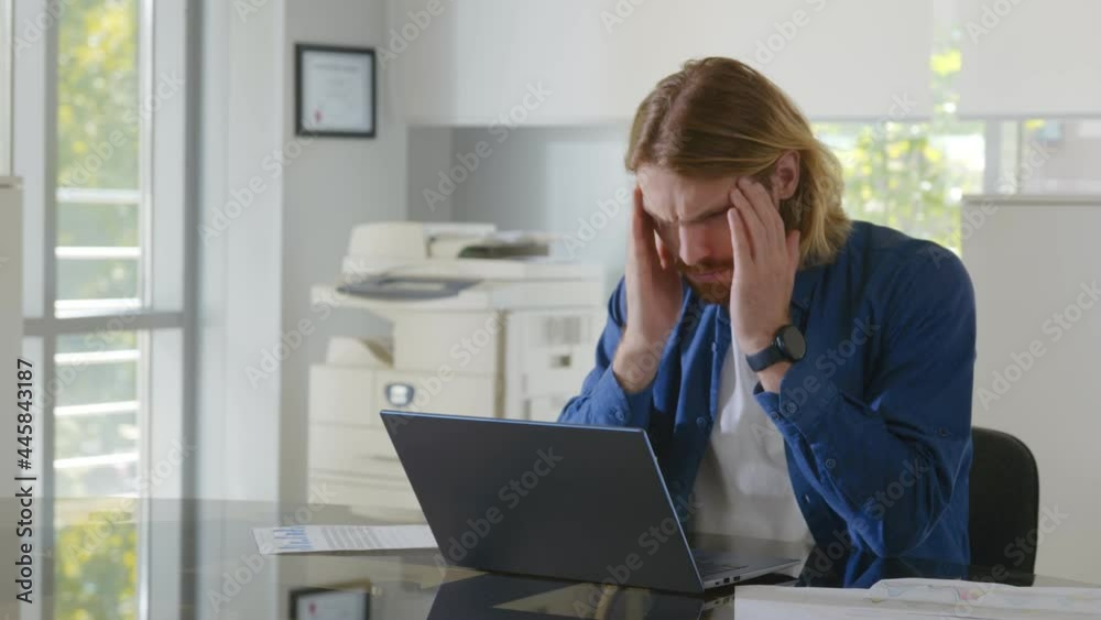Tired young businessman feeling stressed and overwhelmed sitting in modern office with laptop Tired young businessman feeling stressed and overwhelmed sitting in modern office with laptop
