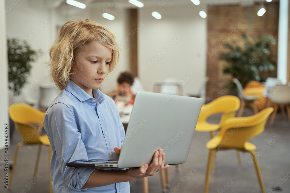 Portrait of calm little boy looking focused while holding and using laptop, standing in a classroom Portrait of calm little boy looking focused while holding and using laptop, standing in a classroom