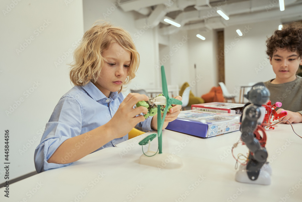 Create things. Caucasian boy examining technical toy while sitting at the table together with other kids, having engineering class