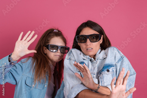Modern mom and daughter in denim jackets on terracotta background in 3d cinema glasses watching horror movie together  scared afraid face
