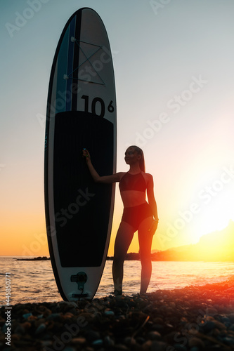 Surfing. Silhouette of a young slender woman holding a sup board. In the background, the ocean and the sunset. Summer vacation at the sea