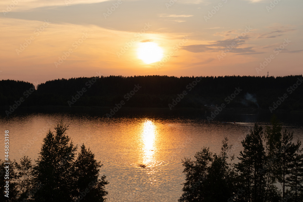 Lake or river in the light of sunset among trees, reflection of sun in the water