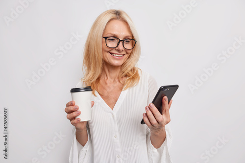 Glad European woman drinks coffee from takeaway cup holds smartphone uses free internet connection during break smiles gently wears spectacles and stylish blouse isolated over white background