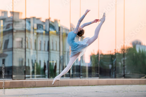 young ballerina in a white leotard is dancing on pointe shoes against the backdrop of the reflection of sunset in the city, frozen in jump