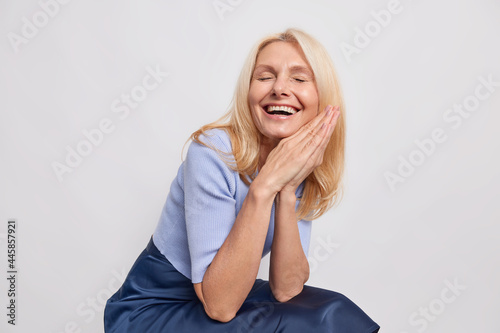 Overjoyed blonde fifty years old woman smiles broadly keeps palms pressed together expresses positive authentic emotions laughs at something dressed in stylish clothes isolated over white background