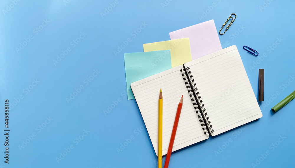 Top view flat lay of blank page notebook in a line with colored pencils, paper clips and pencil caps on blue background Top view flat lay of blank page notebook in a line with colored pencils, paper clips and pencil caps on blue background