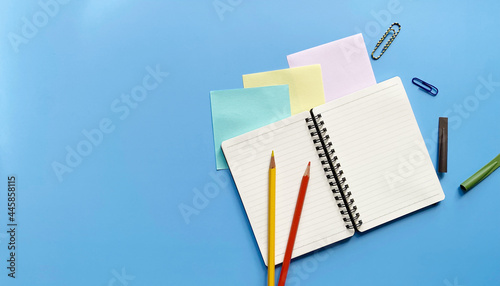 Top view flat lay of blank page notebook in a line with colored pencils, paper clips and pencil caps on blue background