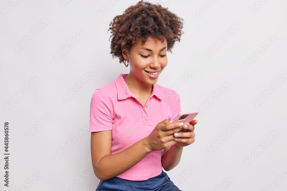 Smiling good looking woman with curly hair uses mobile phone for sending text messages and scrolling newsfeed dressed in casual pink t shirt isolated over white background. Technology concept
