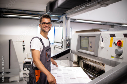 Portrait of smiling caucasian operator standing by paper cutting machine in printing factory.
