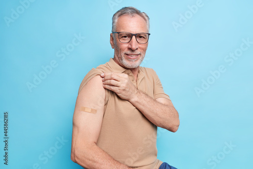 Old man with grey hair shows vaccinated arm motivates to vaccinate against coronavirus to stop epidemic cares about health in his age wears brown t shirt spectacles isolated over blue background photo