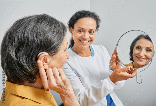 Mature woman with BTE hearing aid looks at himself in mirror held by smiling audiologist, and tries on hearing device. Stylish hearing device