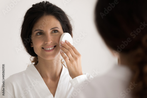 Working on perfect skin. Young hispanic female stand by mirror using extra delicate facial sponge to apply lotion enjoy soft silky touch. Millennial woman make tender face exfoliation by special tool