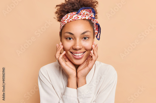 Fotomurale Portrait of lovely dark skinned woman touches face gently smiles broadly has perfect white teeth wears headband and shirt looks happily at camera isolated over beige background