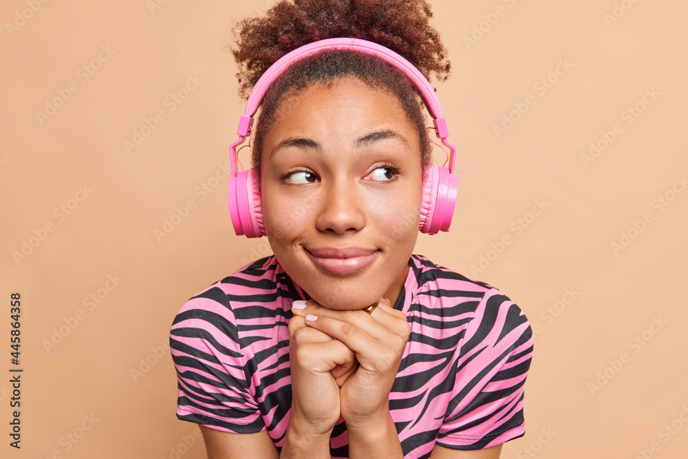 Close up portrait of thoughtful pleased dark skinned woman keeps hands under chin wears headphones on ears listens audio track or tutorial lecture concentrated aside isolated over beige wall Close up portrait of thoughtful pleased dark skinned woman keeps hands under chin wears headphones on ears listens audio track or tutorial lecture concentrated aside isolated over beige wall