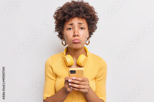 Upset African American woman purses lips looks sadly at camera gets unpleasant message or notification on mobile phone dressed casually uses stereo wireless headphones isolated on white wall