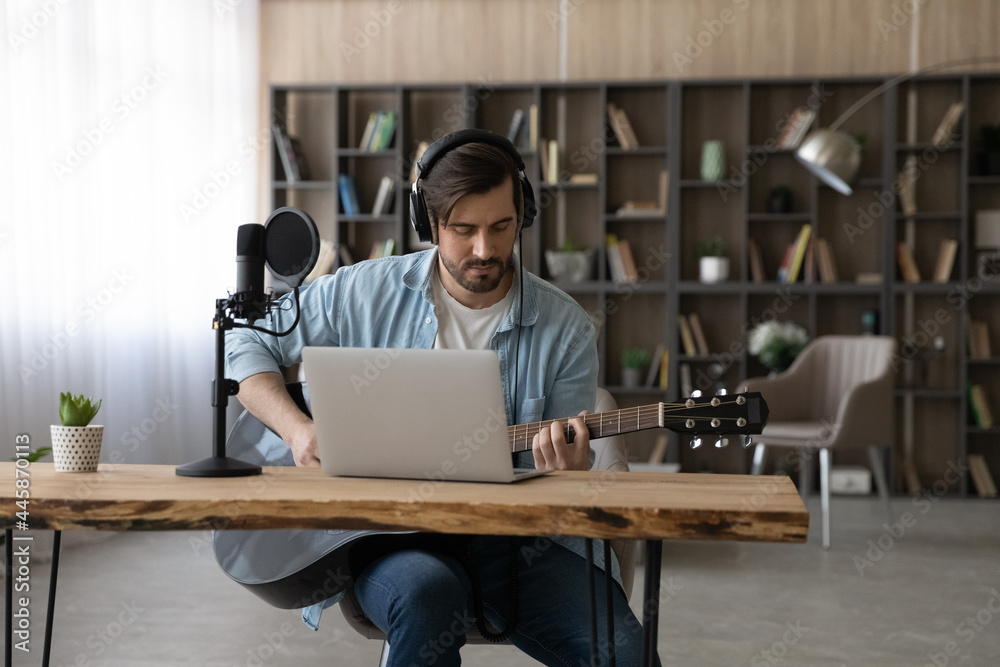 Focused young handsome man wearing headphones, sitting at table with stand microphone and computer playing guitar, watching online music class, improving creative skills distantly at home studio. Focused young handsome man wearing headphones, sitting at table with stand microphone and computer playing guitar, watching online music class, improving creative skills distantly at home studio.